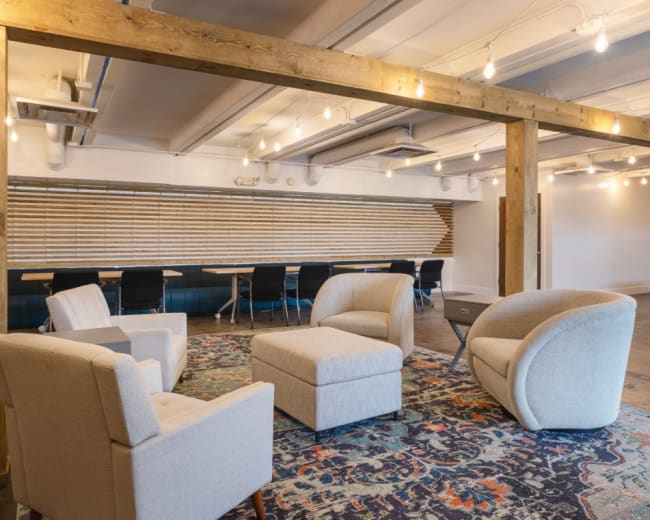 The image shows a modern office space with a seating area featuring two armchairs and an ottoman on a patterned rug, alongside a long table and black chairs in the background.