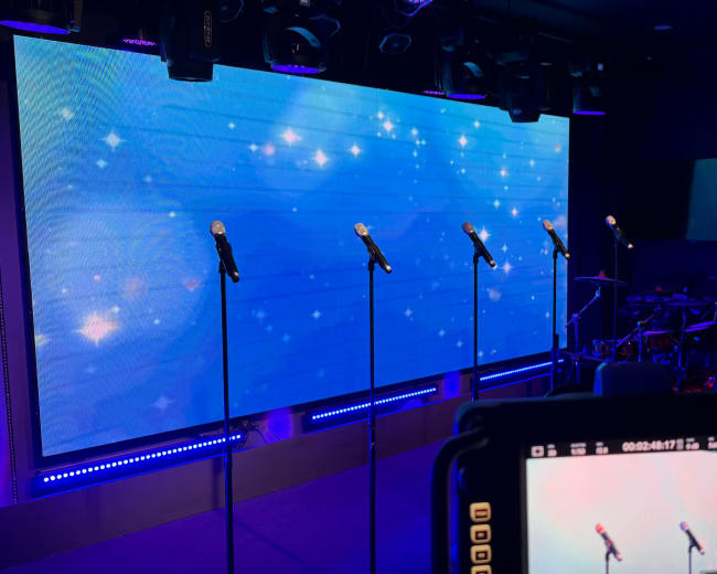 A stage setup features several microphones in front of a large illuminated screen with a blue background, complemented by a drum set and cameras positioned for recording.