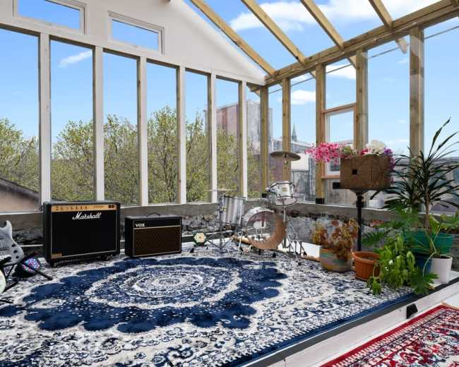 A sunlit music room features electric guitars, amplifiers, and a drum set arranged around a decorative rug, with large windows offering a view of trees outside.