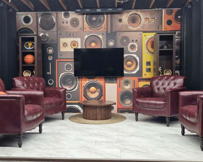 The image shows a room with two red leather armchairs facing a television on a wall decorated with various vintage speakers and sound equipment.