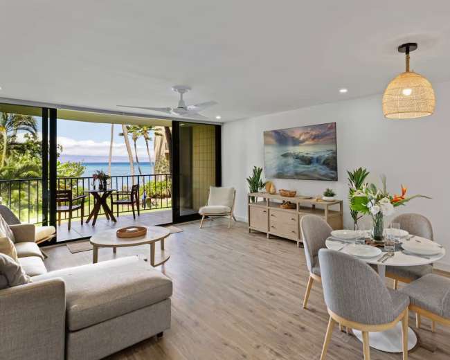 The image shows a modern living and dining area with a view of the ocean, featuring a sofa, dining table, and large windows that open to a balcony.