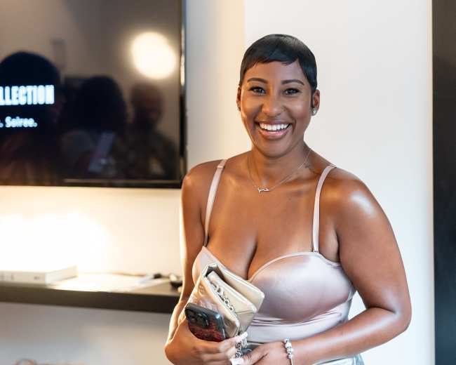 A woman with short hair and a light-colored dress smiles while holding a clutch bag in a modern indoor setting.