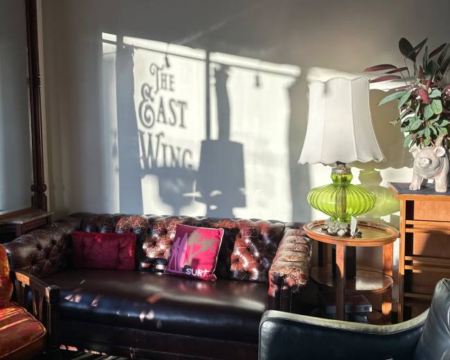 A well-furnished living space features a leather couch with colorful pillows, a lamp, and a decorative plant, while shadows create the text "THE EAST WING" on the wall.