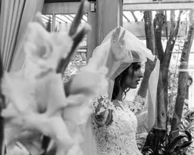A bride adjusts her veil while standing beside a floral arrangement in a glass-enclosed setting.