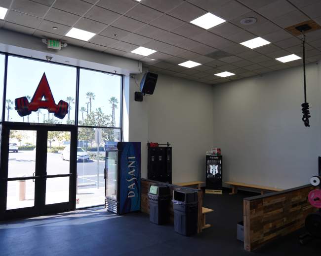 The image shows the interior of a gym with a large logo above the entrance, including racks for weights and benches along the walls.