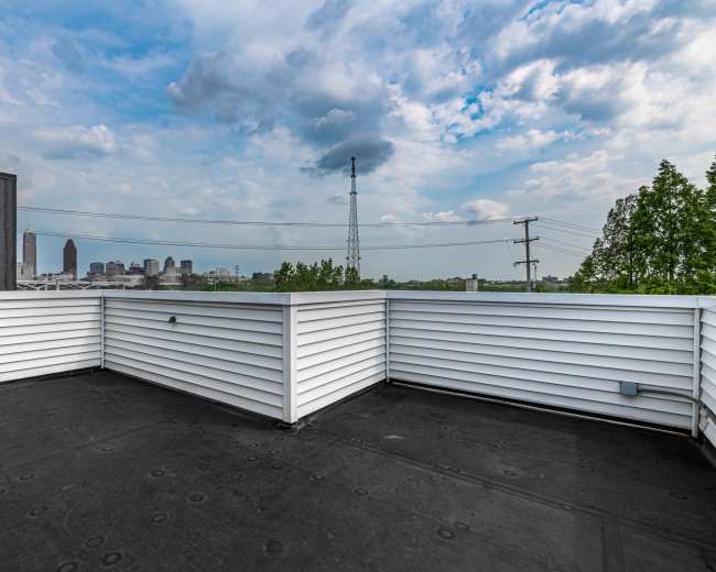 The image shows a rooftop terrace with white railings overlooking a city skyline under a partly cloudy sky.