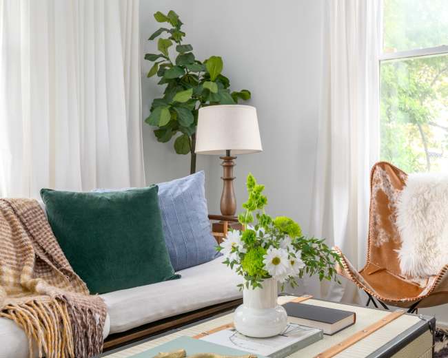 The image shows a cozy living room with a sofa adorned with pillows, a decorative table featuring a vase of flowers, and a butterfly chair beside a lamp and a large window.