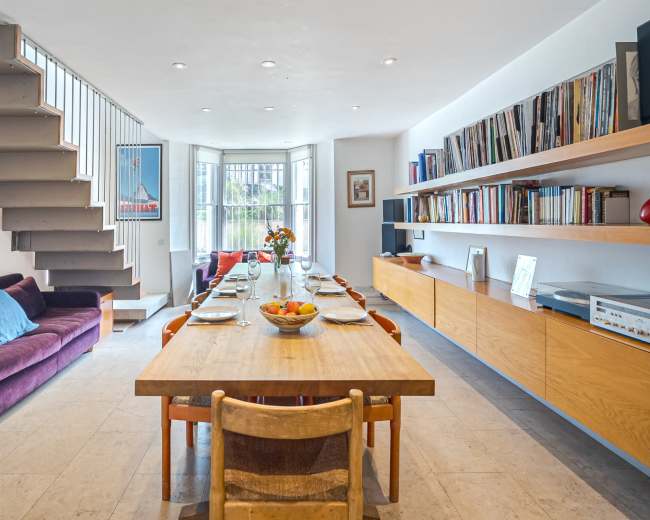 A dining area features a wooden table set for eight, surrounded by chairs, with a sofa and bookshelves along the walls and stairs leading to the upper level.