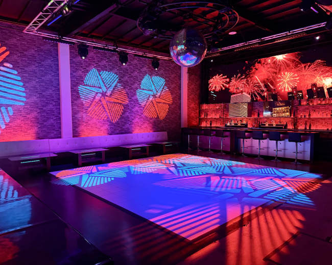 The image shows a vibrant nightclub interior with colorful lighting effects on the walls and dance floor, along with a bar area in the background.