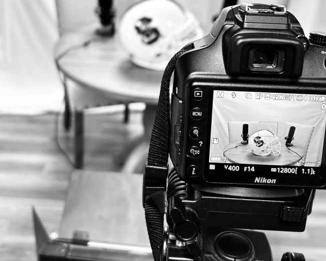 A camera is positioned in front of a table with a football helmet and two microphones, set against a white backdrop.