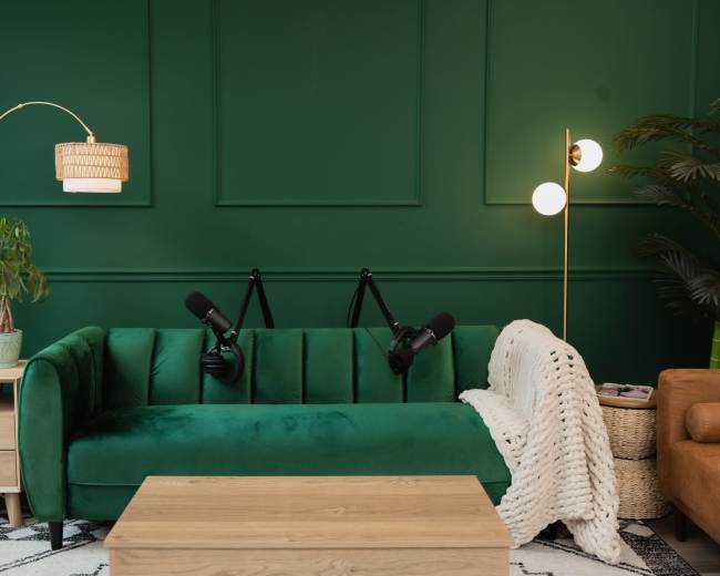 The image shows a green velvet couch with two microphones mounted on stands, surrounded by a wooden coffee table, a woven blanket, a potted plant, and decorative lamps against a dark green wall.