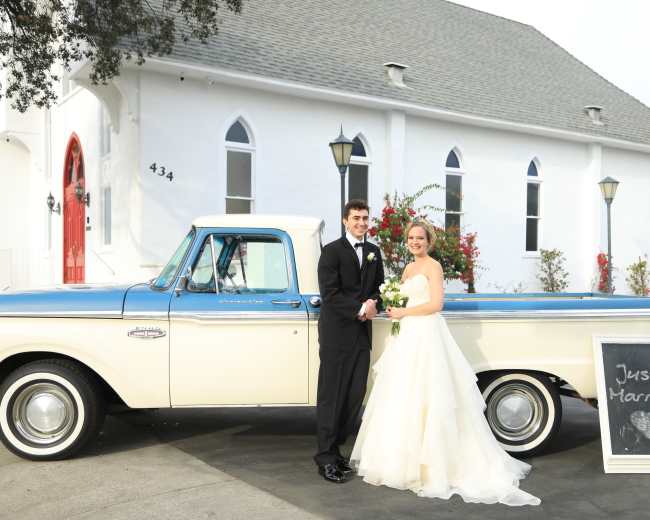 A newly married couple stands beside a vintage pickup truck decorated with a "Just Married" sign outside a white church.