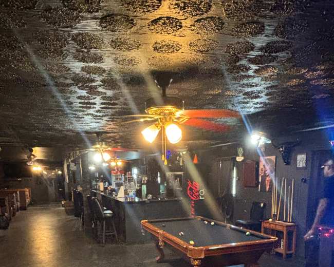A dimly lit room features a billiard table surrounded by chairs and bar stools, with a ceiling fan overhead.