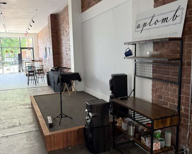 The image depicts an interior space featuring a small stage with a microphone, a sound system on a black shelf, and a sign that reads "aplomb," all within a brick-walled venue.