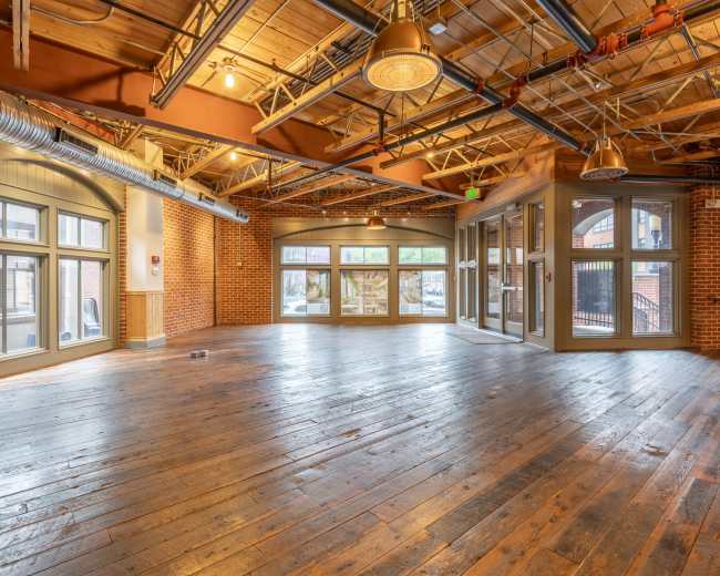 The image shows an empty room with wooden floors, exposed beams, and large windows that provide natural light.