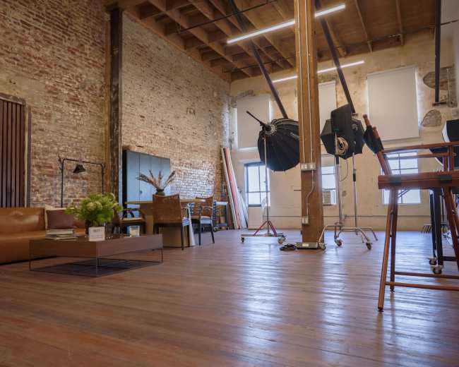 The image shows a spacious, industrial-style room with exposed brick walls, wooden beams, and a mix of furniture including a leather sofa, a coffee table, and photography equipment.