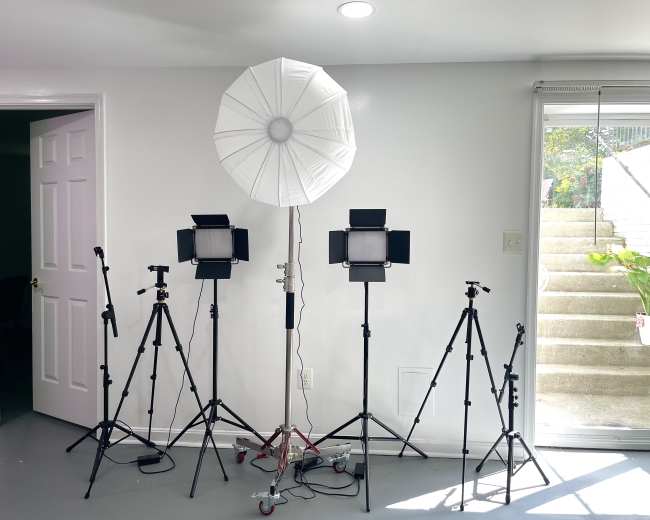 The image shows a photography setup featuring a large white umbrella softbox and several tripods with lights arranged in a room near a glass sliding door.