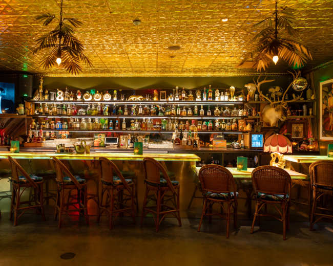 The image shows a vibrant bar interior with a long counter, numerous bottles displayed on shelves, and ambient lighting, including ceiling decorations.