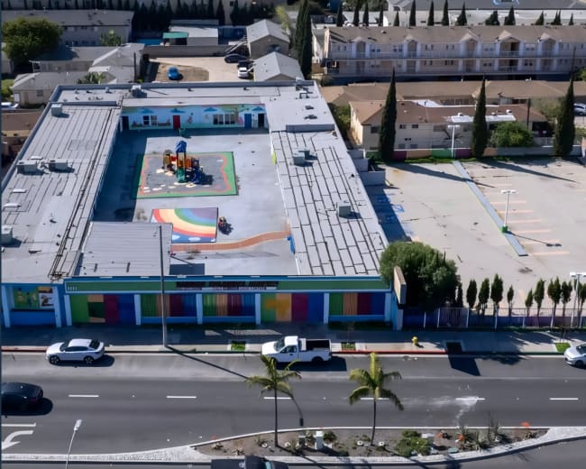The image shows an aerial view of a colorful building with a playground featuring a rainbow design, surrounded by a parking area and several trees.