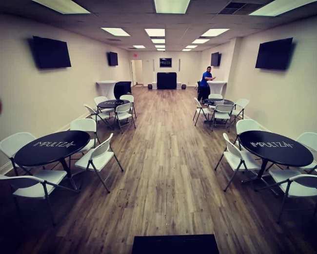 A spacious, well-lit event room with several round black tables and white chairs arranged throughout, along with two wall-mounted televisions and a small stage at the back.