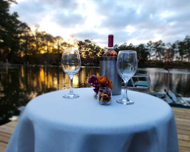 A table set with two wine glasses, a bottle of wine in a cooler, and a small vase of flowers overlooks a tranquil lake at sunset.