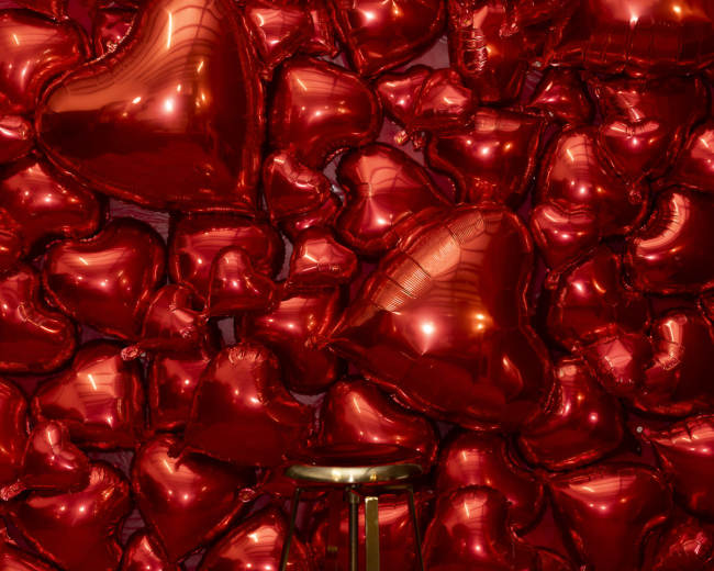 A gold stool is positioned in front of a wall covered in red heart-shaped balloons.