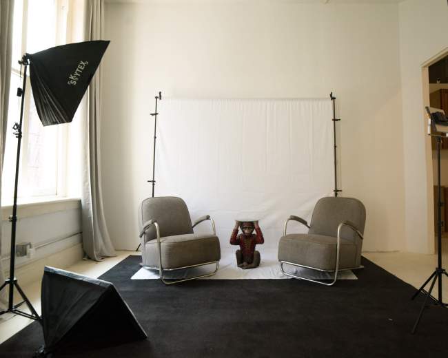 A minimal photography studio setup features two grey chairs, a black backdrop, a white screen, and lighting equipment.