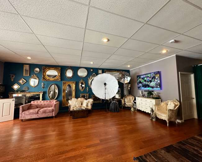 The scene shows a spacious room with a variety of mirrors arranged on one wall, a photography setup with a large softbox, and a mix of vintage and contemporary furniture.