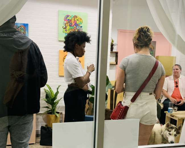 A group of people are gathered inside a brightly lit room with plants, while a dog sits on the floor near them.