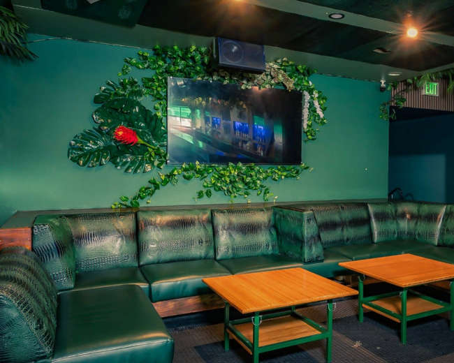 The image shows a spacious lounge area with green leather seating, two wooden tables, and a wall decorated with plants and a television.