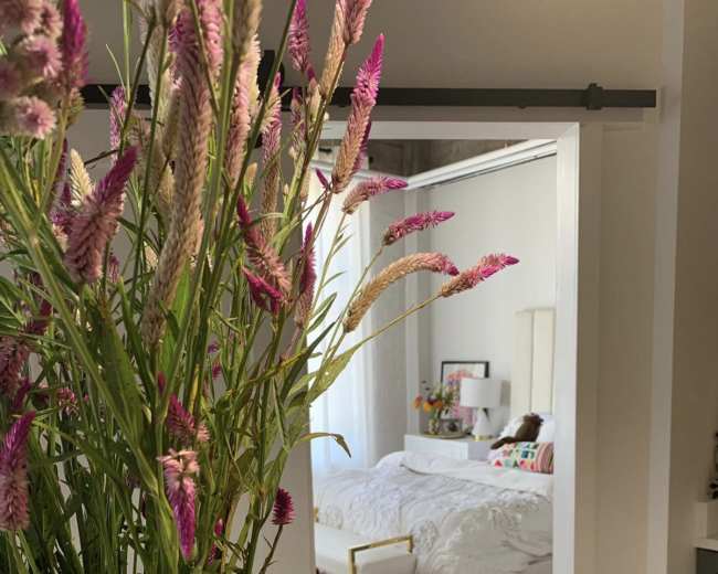 A bouquet of pink flowers in a vase is placed in the foreground, with a glimpse of a bedroom featuring a bed and colorful decor in the background.
