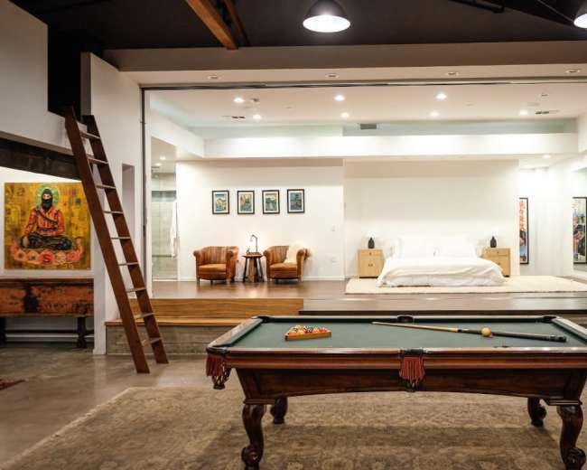 The image features a spacious loft with a pool table in the foreground and a minimalist bedroom area visible in the background.