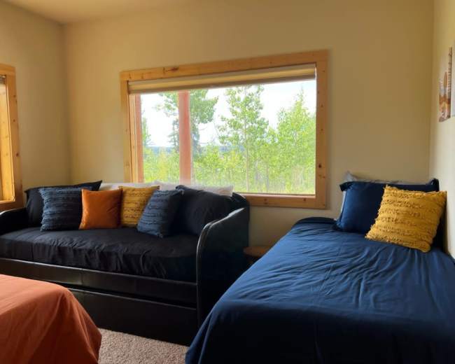 The image shows a cozy bedroom featuring two beds, one with a navy-blue cover and the other with black, both positioned near a large window that offers a view of trees outside.