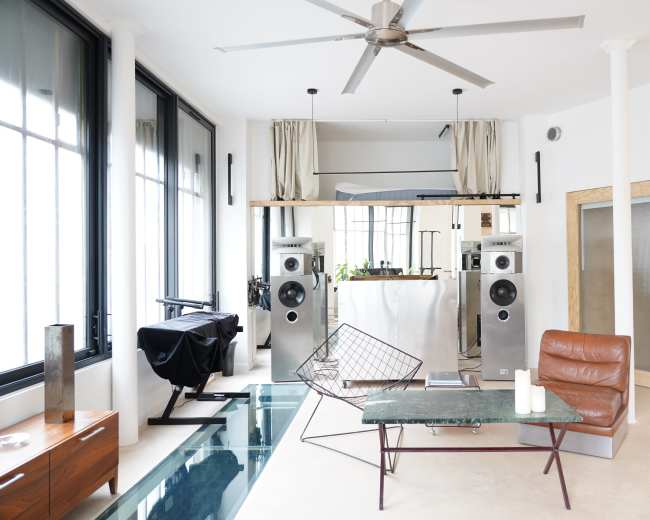 A modern living room features large windows, a sleek kitchen in the background, white walls, a green marble coffee table, and two tall speakers flanking the space.