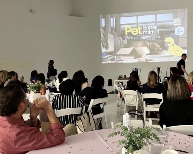 A presentation is being held in a well-lit room with an audience seated at round tables, while a large screen displays a promotional video for a pet supermarket.