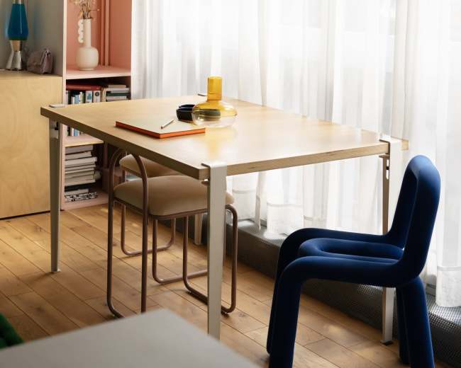 A modern room features a wooden table with a round black plate and a yellow glass, complemented by a blue chair and natural light filtering through sheer curtains.