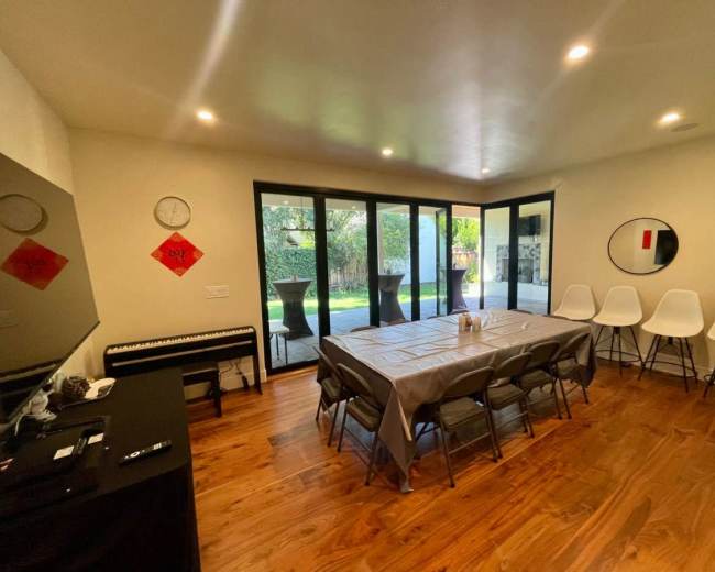 The interior of a spacious room features a long table with chairs arranged around it, a piano against one wall, and large glass doors leading to an outdoor area.