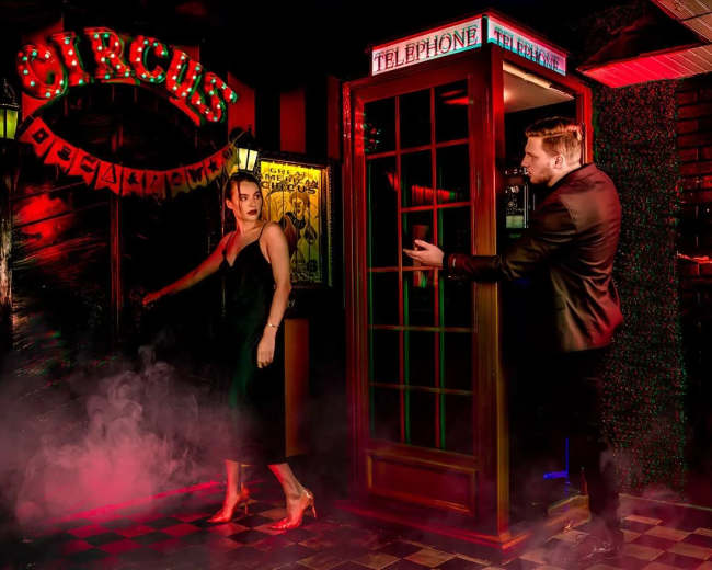 A woman in a black dress exits a bar illuminated by neon signs, while a man in a suit gestures from a nearby telephone booth.