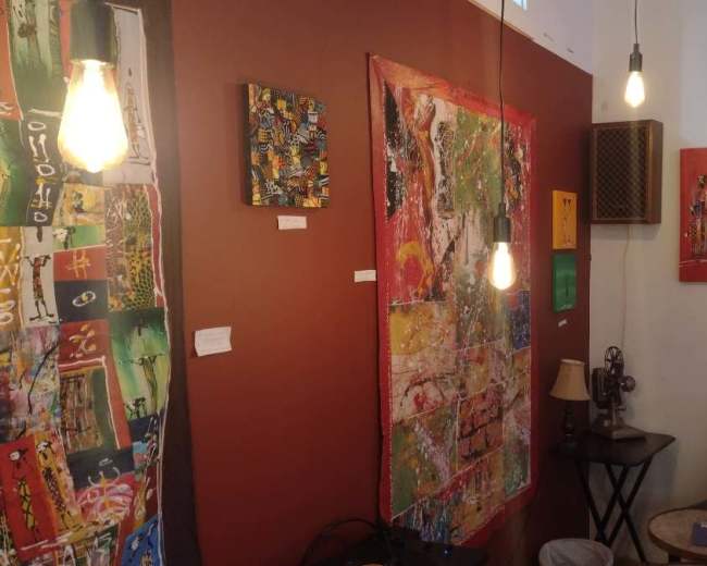The image shows a cozy art space featuring colorful abstract paintings on the walls, with a small seating area and hanging light bulbs.