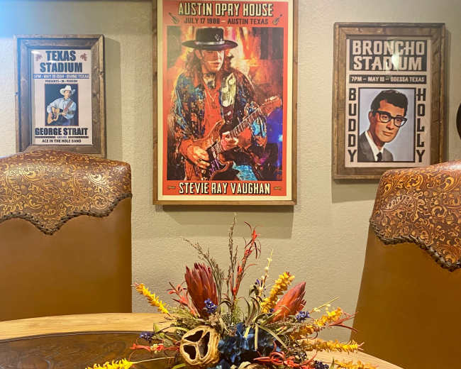 The image shows a dining area with a circular wooden table, decorated with a floral arrangement, and walls featuring framed posters of music venues and artists, including an Austin Opry House poster of Stevie Ray Vaughan.