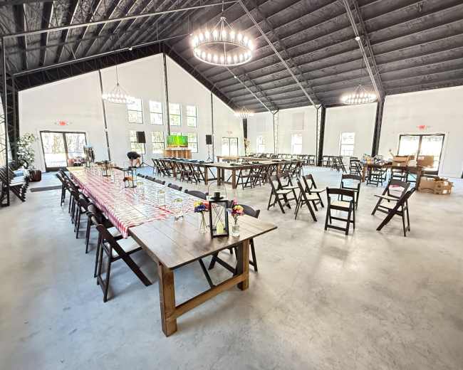 The image shows an indoor event space with wooden tables and chairs arranged for a gathering, featuring high ceilings and large windows providing natural light.
