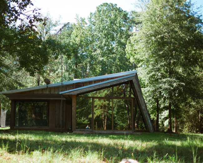 A modern house with large glass windows and a slanted roof is situated among trees in a grassy area.