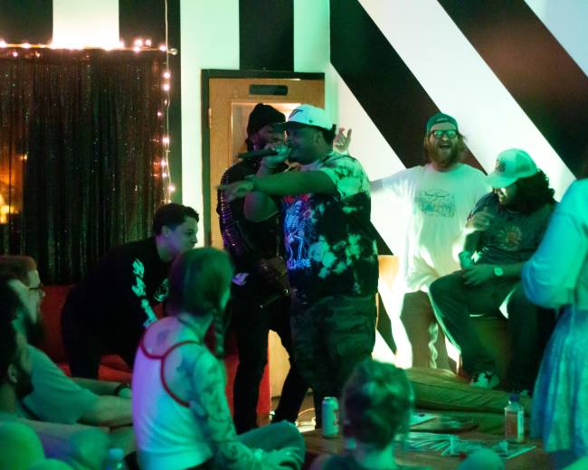 A group of performers engages with the audience during a lively event held in a room decorated with black and white striped walls and ambient lighting.