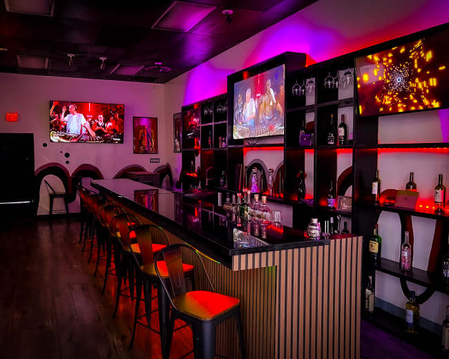 A modern bar area with a polished counter, high stools, and illuminated shelves displaying various bottles, flanked by large screens showing videos.