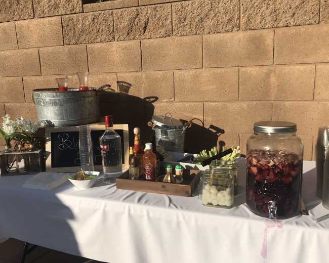 A table set up with various bottles of alcohol, mixers, and garnishes, with a chalkboard sign displaying the word "Beverages" against a textured wall.