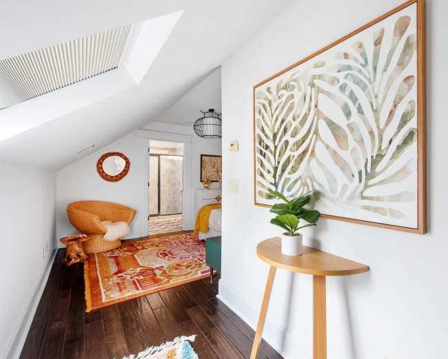 The image shows a bright, small hallway with a decorative patterned rug, a piece of wall art featuring plant motifs, and a small wooden table with a potted plant.