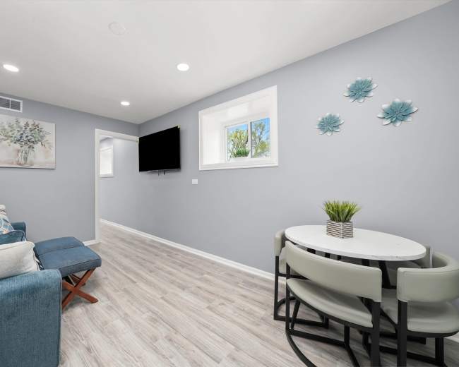 The image shows a modern, light-colored living space with a blue sofa, a wall-mounted TV, and a small dining table with chairs.