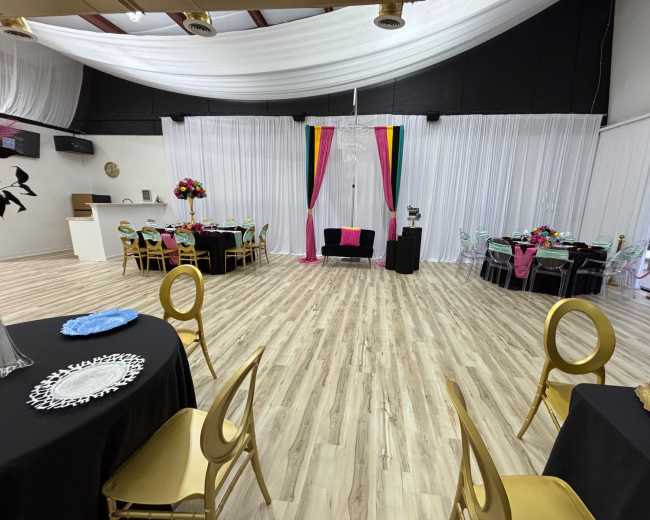 The image shows a decorated event space with tables set for dining, colorful floral centerpieces, and curtains forming a backdrop.