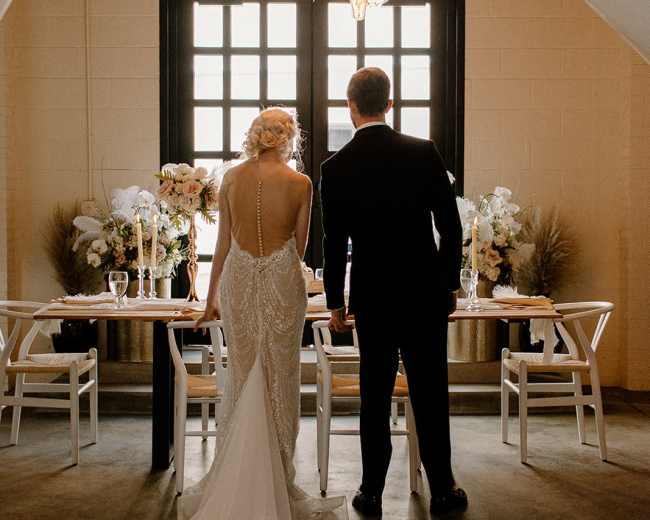 A bride and groom stand together, facing a elegantly set dining table adorned with floral arrangements and candles, under warm hanging lights in a stylish indoor venue.