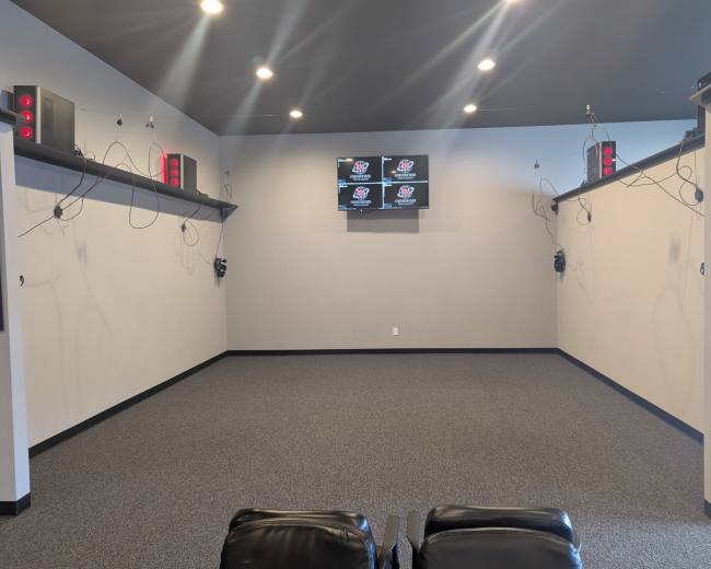 The image shows a spacious room with grey walls, a carpeted floor, and multiple screens mounted on the wall, alongside audio equipment and hanging cables.